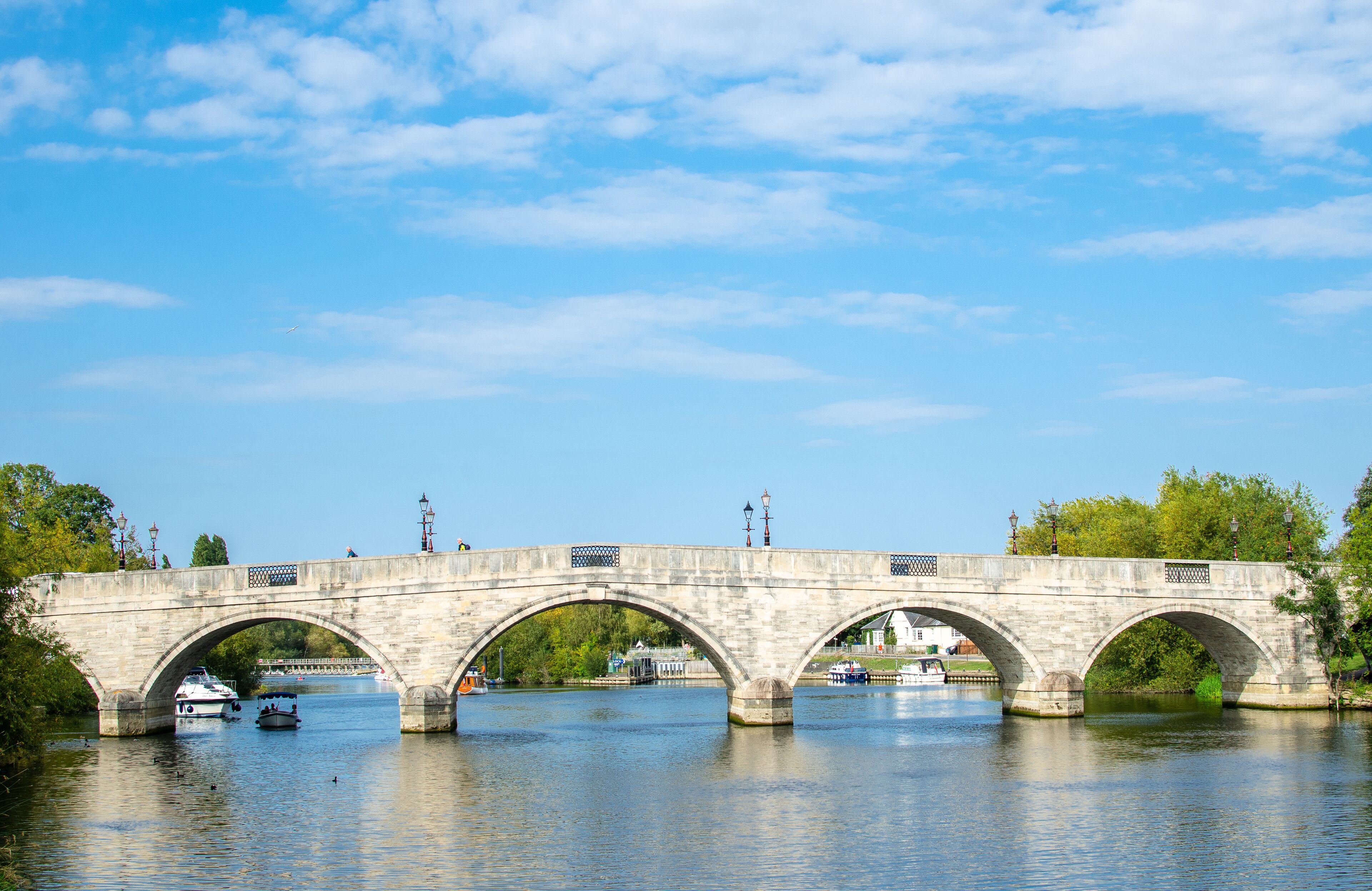 A stone bridge with three arches spanning a river, surrounded by trees and boats.