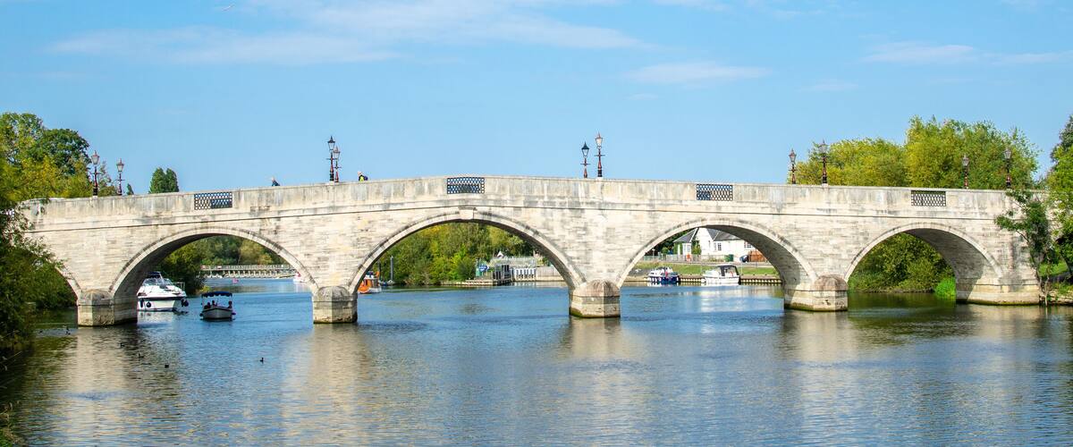 A stone bridge with three arches spanning a river, surrounded by trees and boats.