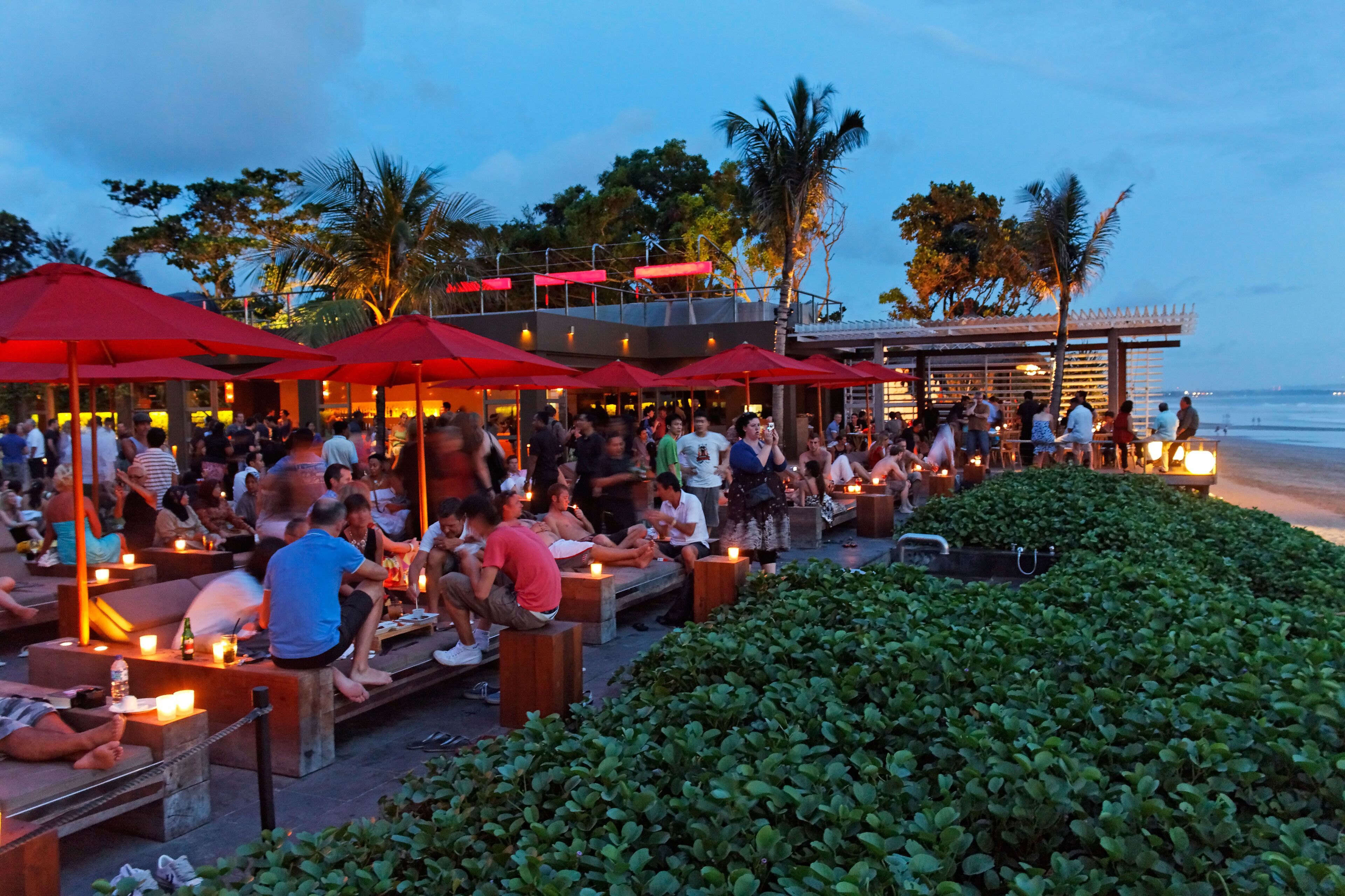 A crowded outdoor bar with red umbrellas and people sitting on benches.