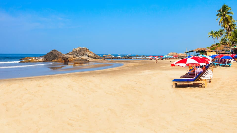 A sandy beach with a rocky outcrop, palm trees, and beach umbrellas.