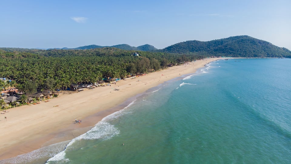 A coastal beach with clear turquoise waters, a sandy shore, and a backdrop of dense green forested hills.