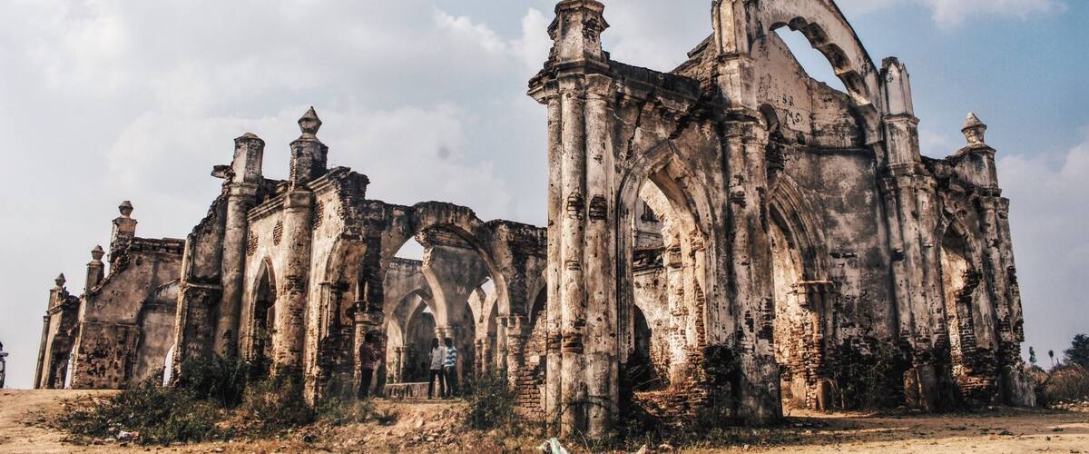 An ancient, weathered stone structure with multiple arches and columns, surrounded by overgrown vegetation.