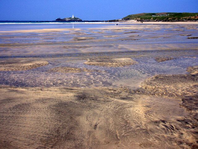 A beach with clear water, a lighthouse, and a grassy hill in the background.