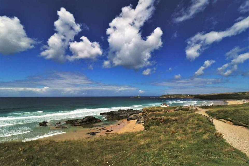 A coastal landscape with a sandy beach, rocky outcrops, and a clear blue sky with scattered clouds.