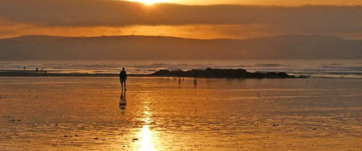Un atardecer sobre una playa, con una persona caminando y una cadena montañosa a lo lejos.