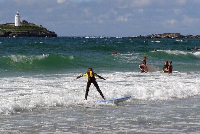A surfer on a wave, a lighthouse, and rocky outcrops in the background.