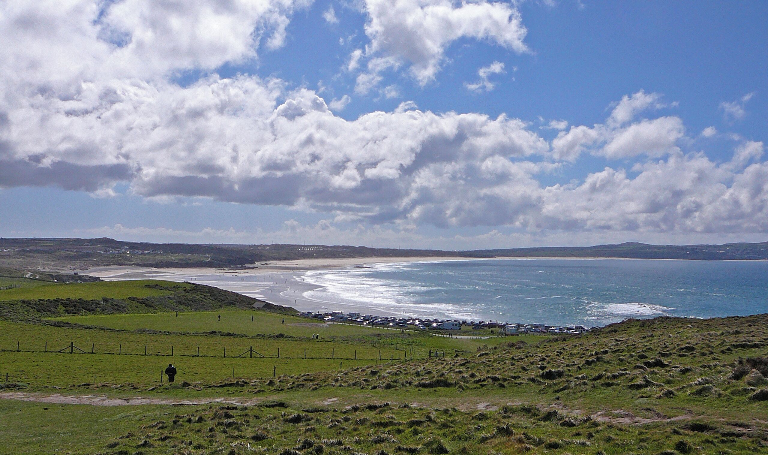 A coastal landscape with a beach, grassy fields, and a cloudy sky.