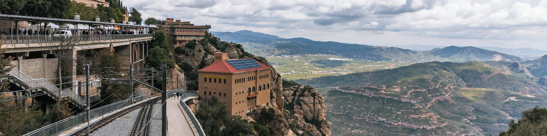 A mountain railway with a pedestrian bridge, a historic building, and a valley view.