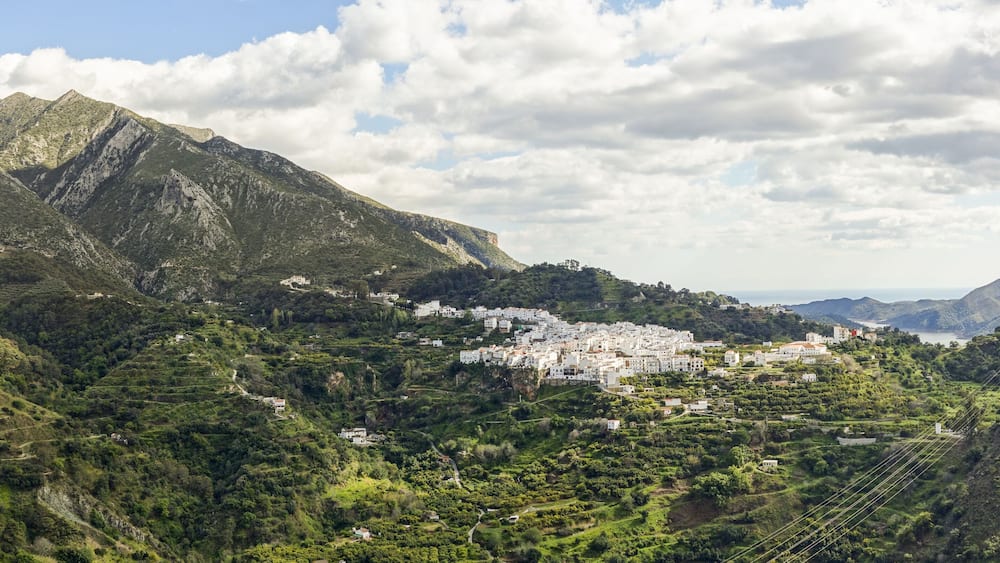A mountainous landscape with terraced fields, a white village, and a coastline in the distance.
