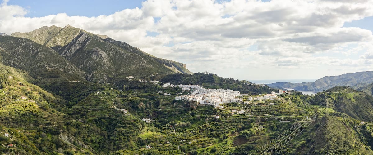 A mountainous landscape with terraced fields, a white village, and a coastline in the distance.