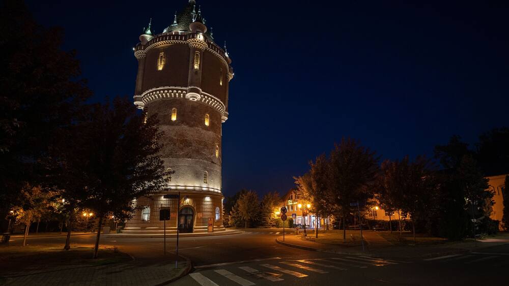 A historic tower with illuminated windows and a clock, surrounded by trees and a paved area.