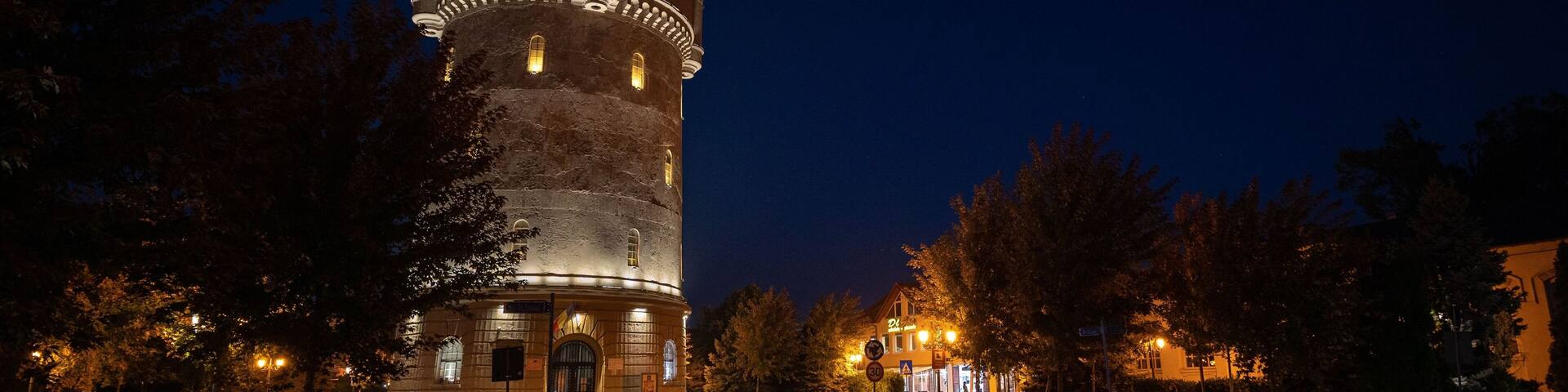 A historic tower with illuminated windows and a clock, surrounded by trees and a paved area.