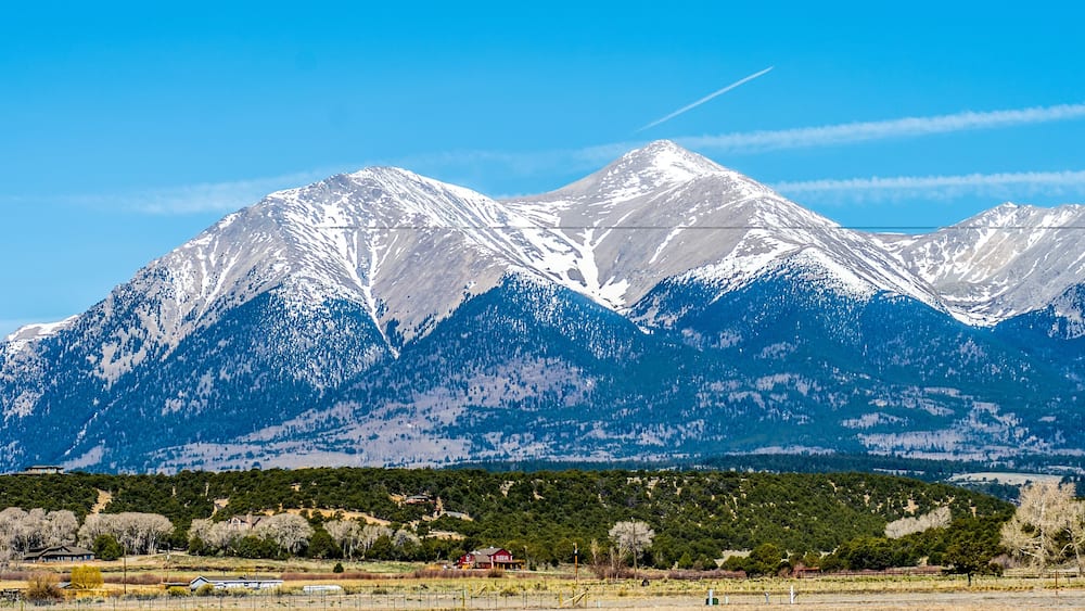 A snow-covered mountain range with a clear blue sky and a few houses in the foreground.