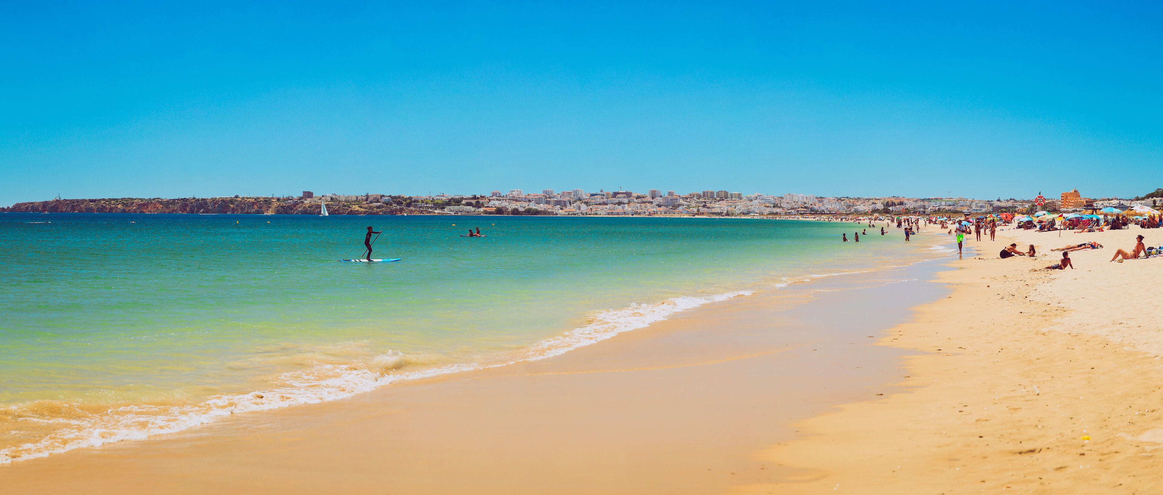 Una playa con aguas turquesas cristalinas, gente tomando el sol y un horizonte urbano a lo lejos.