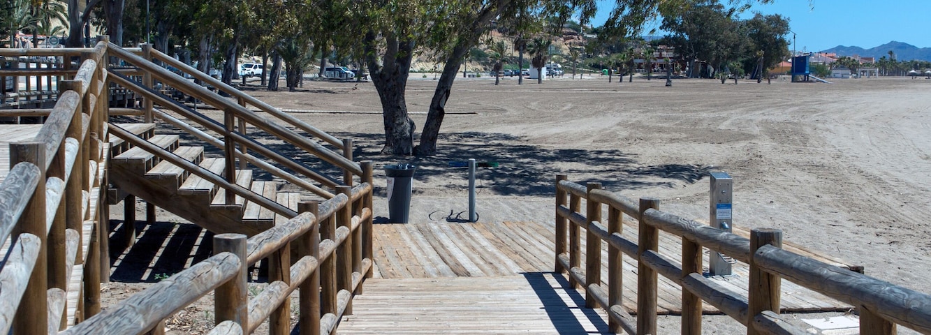 Un muelle de madera que conduce a una zona de playa arenosa, con árboles y cielos despejados.