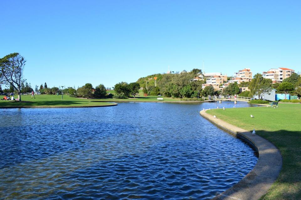 A calm lake with a curved path, green grass, trees, and residential buildings in the background.