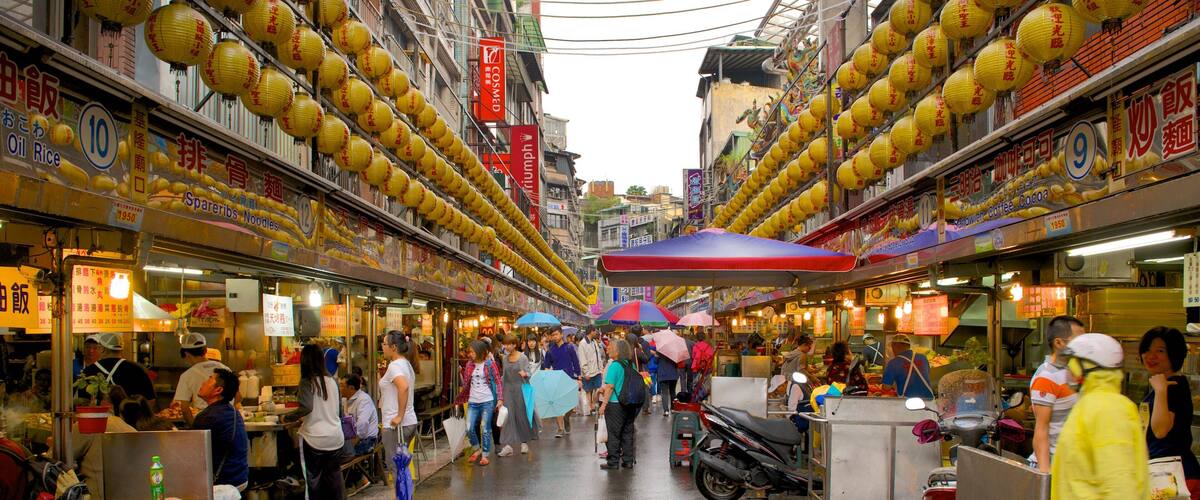 A crowded street market with yellow lanterns, food stalls, and umbrellas.