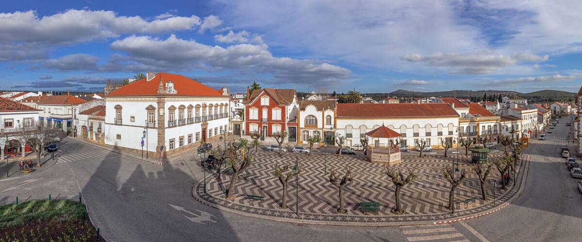 A historic town square with cobblestone streets, traditional buildings, and a central plaza with trees and benches.