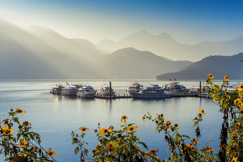 Uma marina com barcos atracados, rodeada por montanhas e águas banhadas pelo sol.