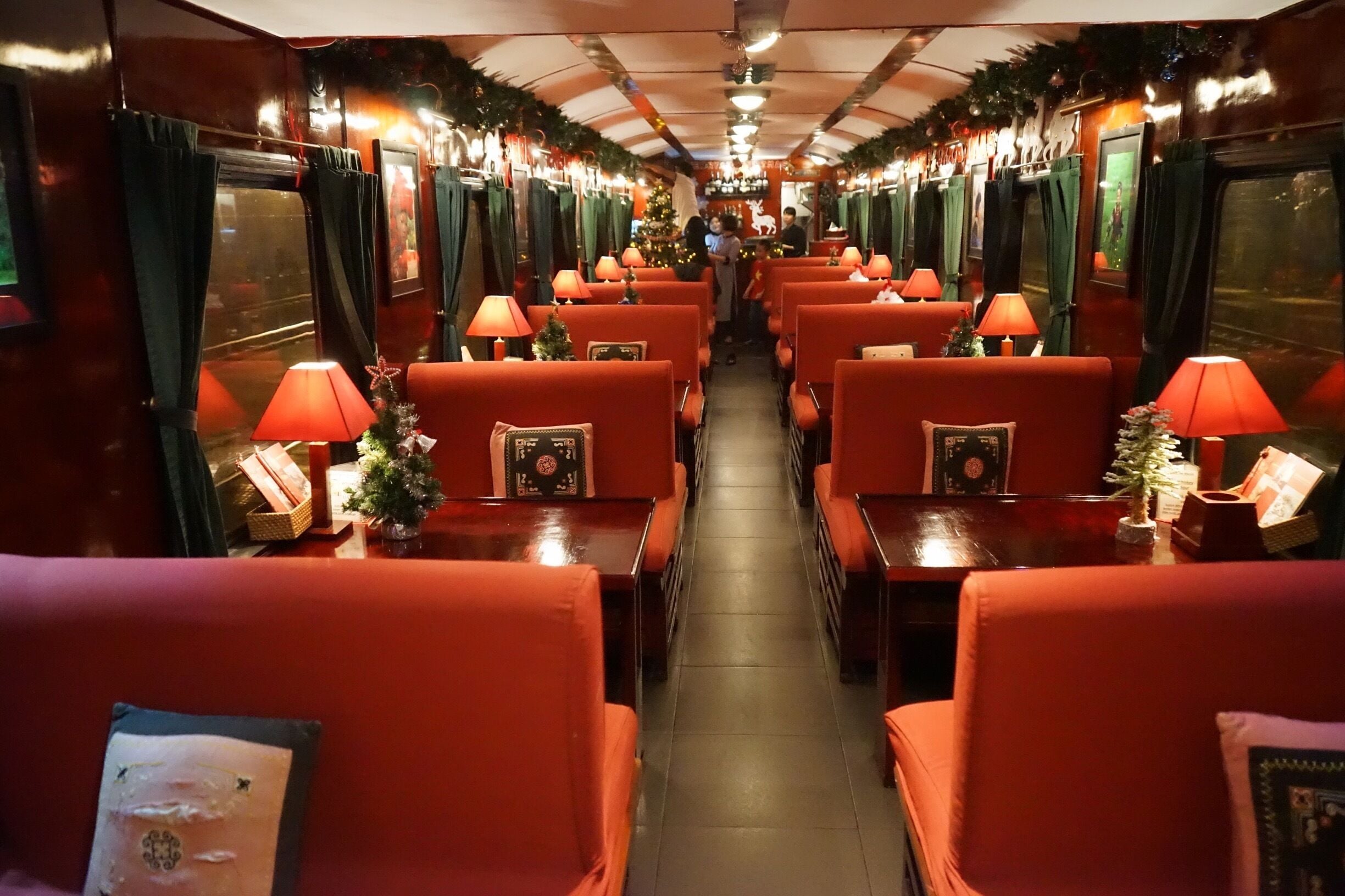 A train interior with red upholstered seats, small tables with lamps, and Christmas decorations.