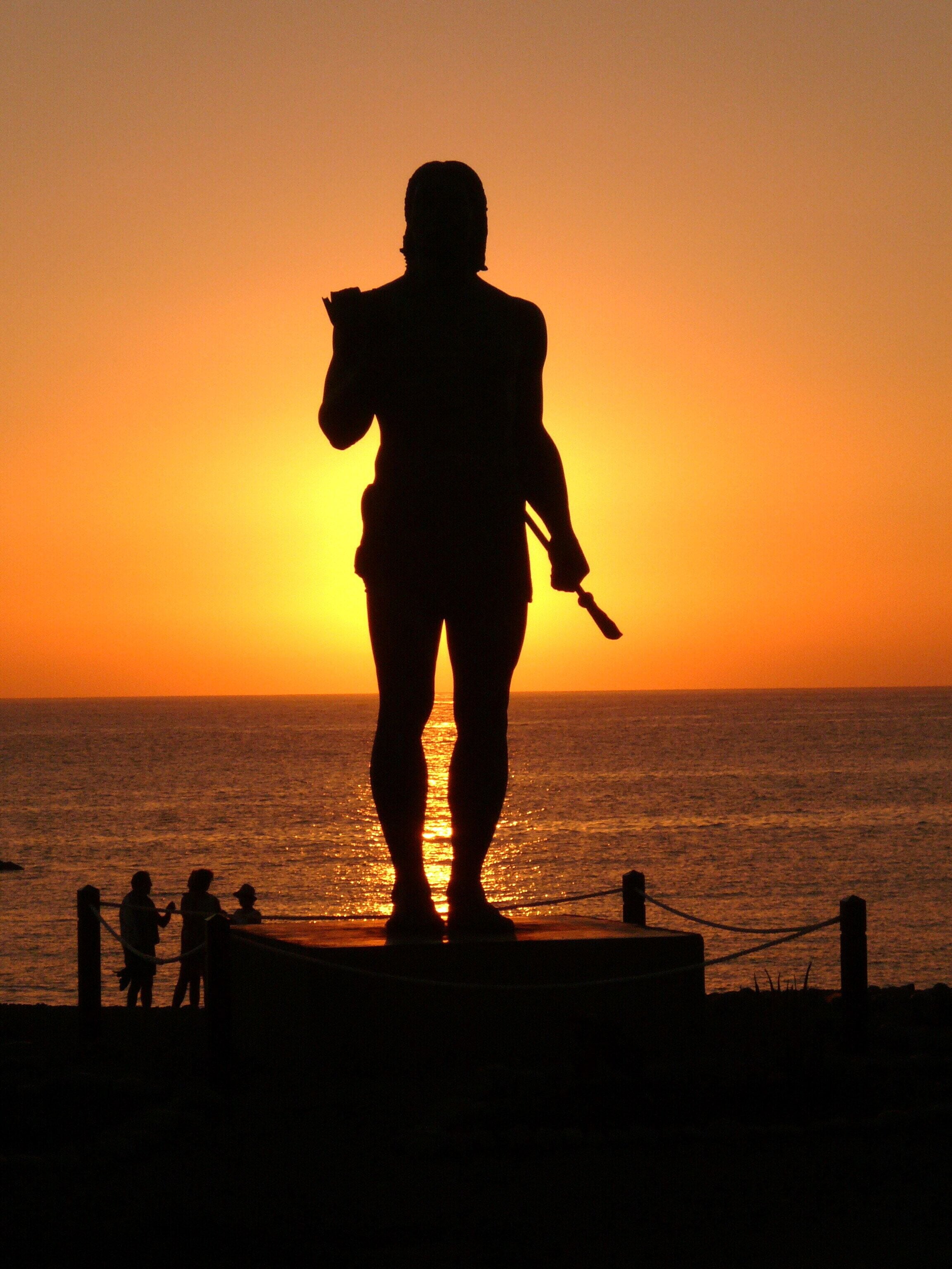 A statue of a person holding a staff, silhouetted against a sunset over the ocean.