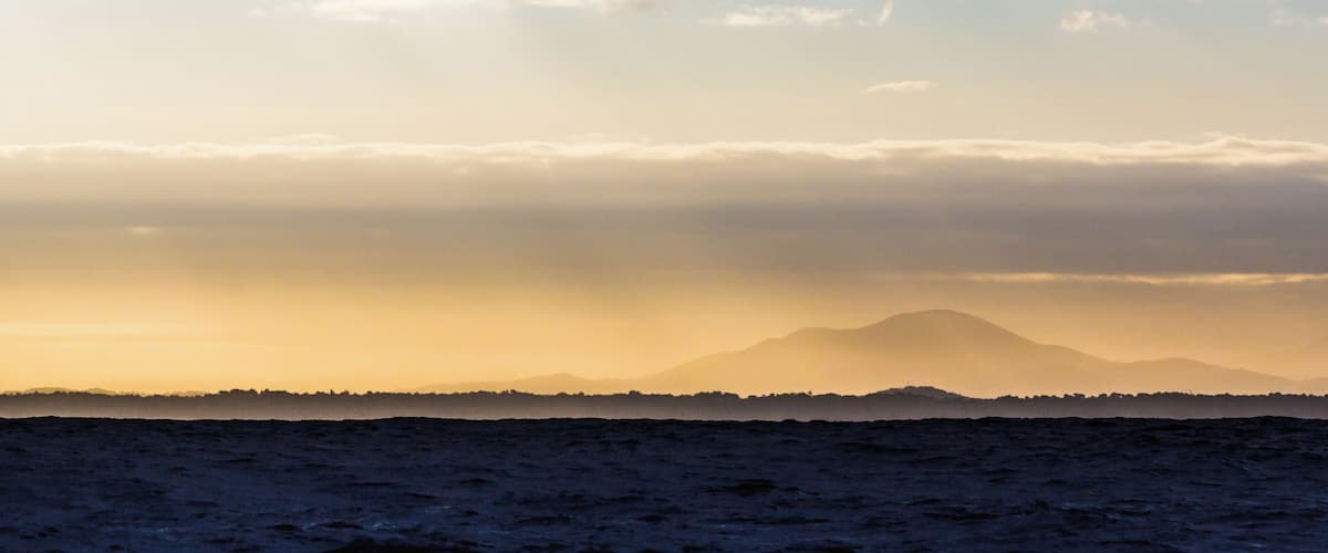 A coastal landscape with a mountain in the distance, ocean waves, and a cloudy sky.