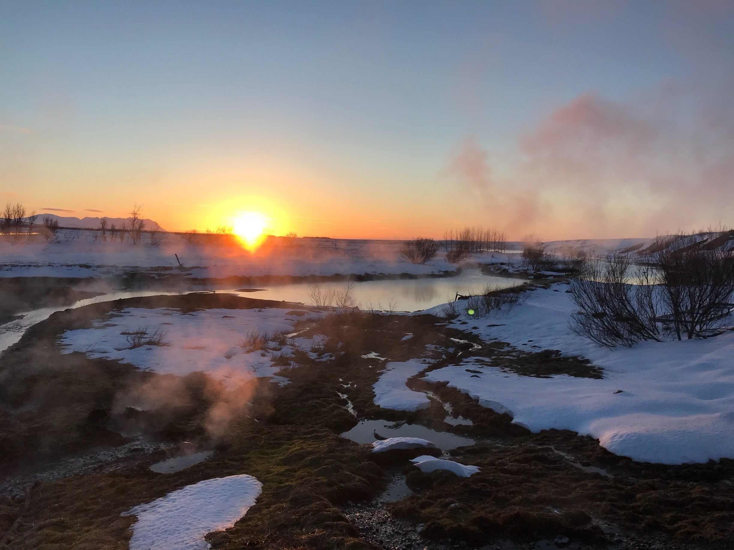 A sunset over a frozen landscape with a body of water and steam rising from the ground.