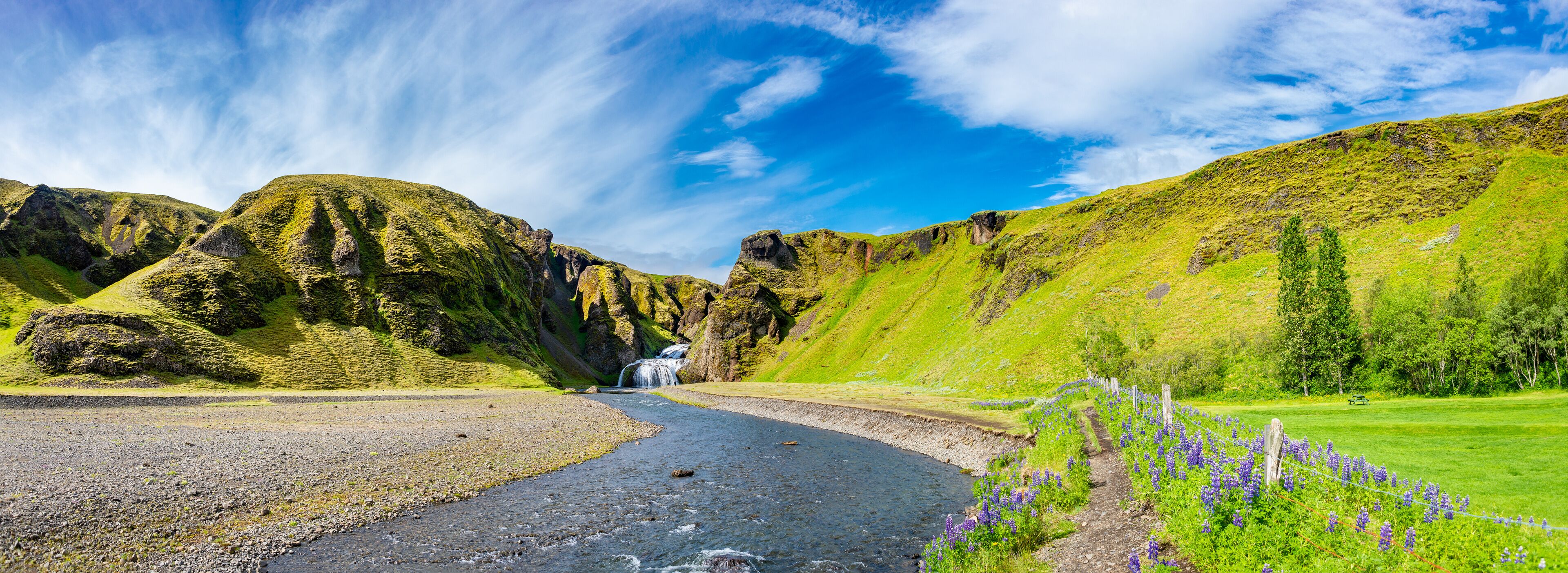 A river flowing through a valley with mountains, a waterfall, and a path with purple flowers.