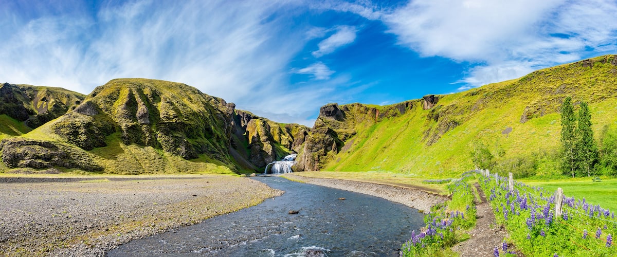 A river flowing through a valley with mountains, a waterfall, and a path with purple flowers.