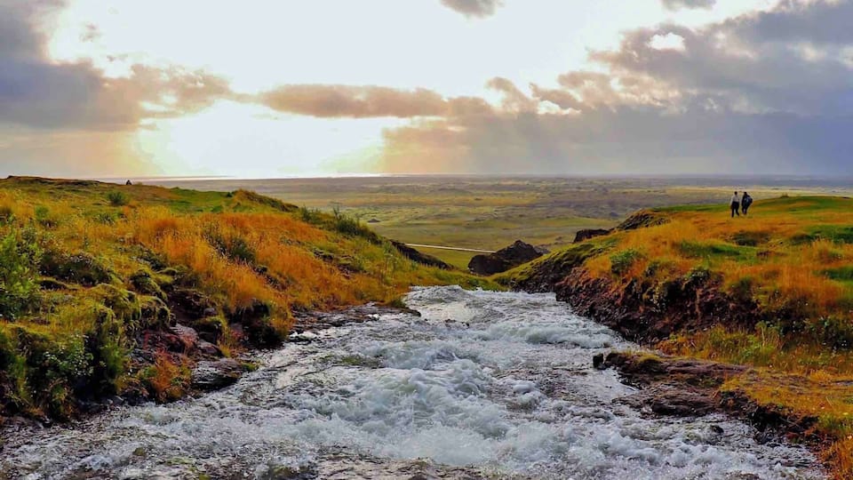 A river flowing through a grassy landscape with two people standing on a hill in the distance.