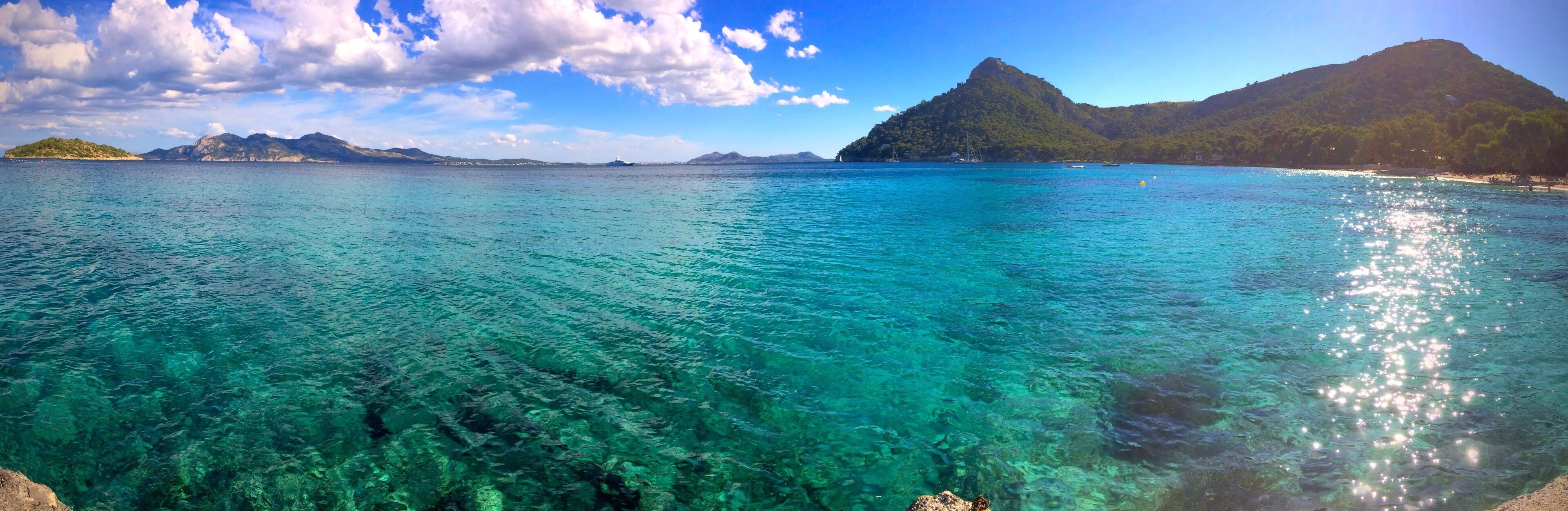 A clear turquoise sea with visible underwater rocks, a small island with a hill, and a mountain in the background.