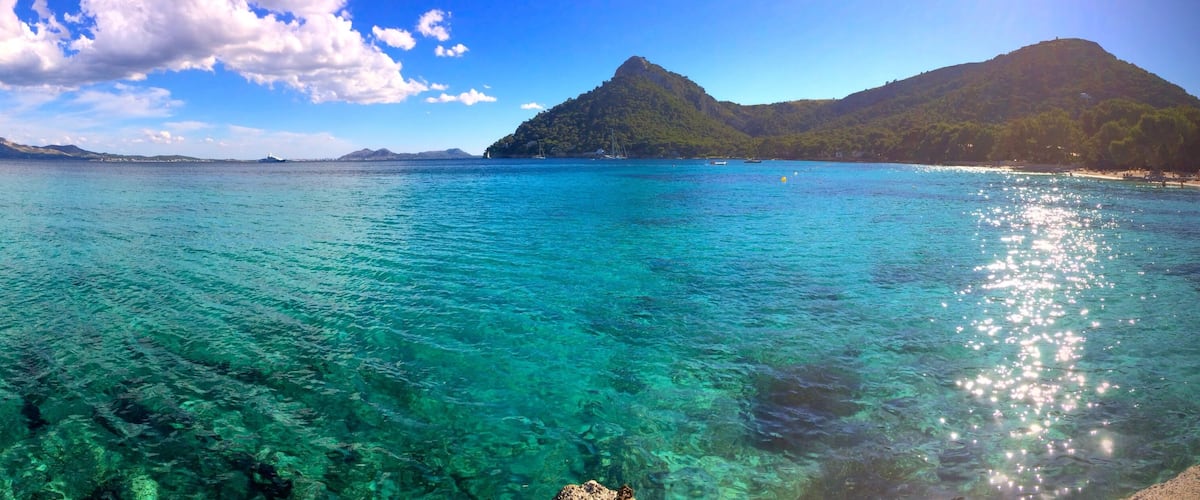 A clear turquoise sea with visible underwater rocks, a small island with a hill, and a mountain in the background.