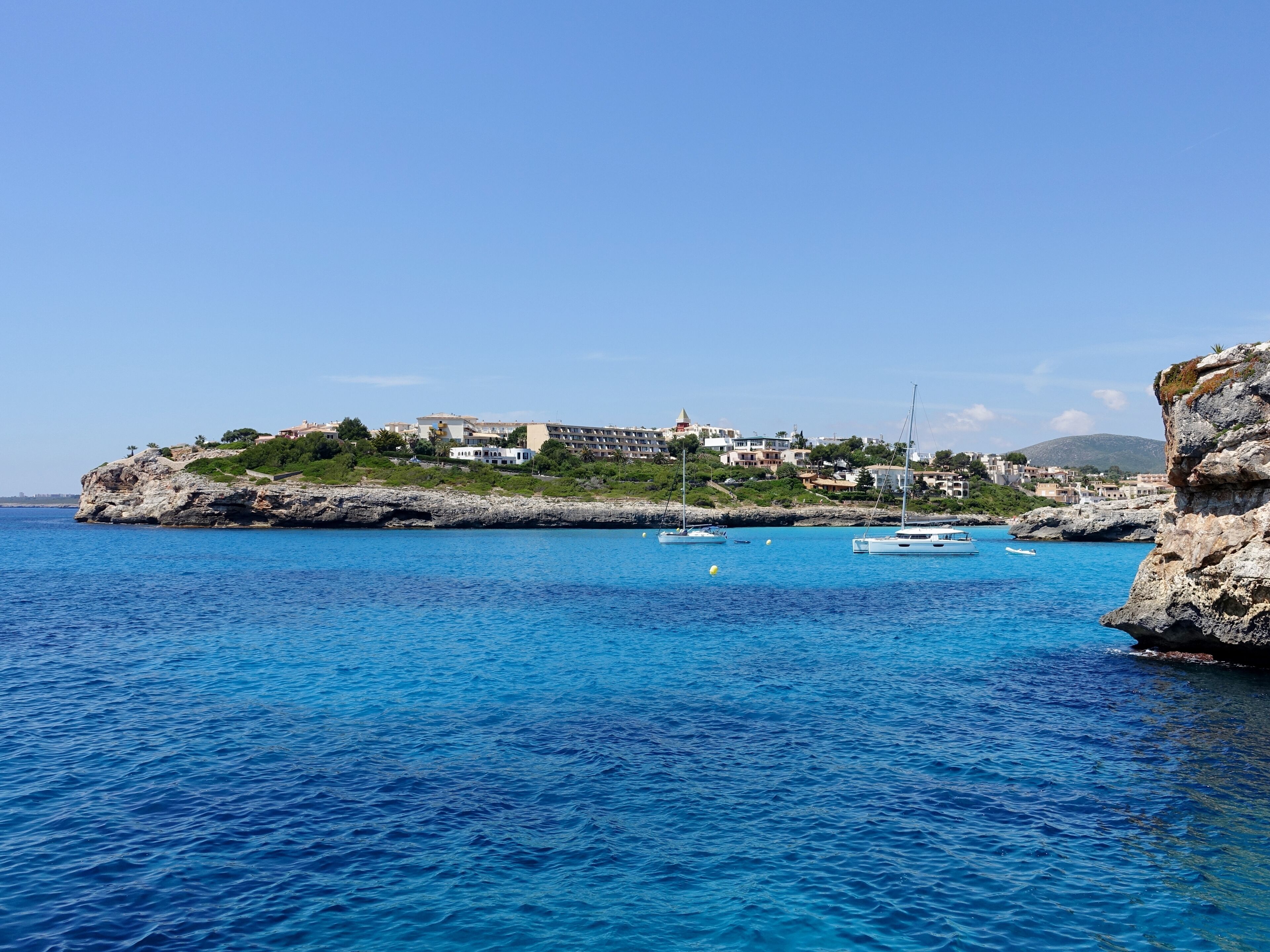 A coastal town with buildings on a rocky outcrop, surrounded by clear blue waters and boats.