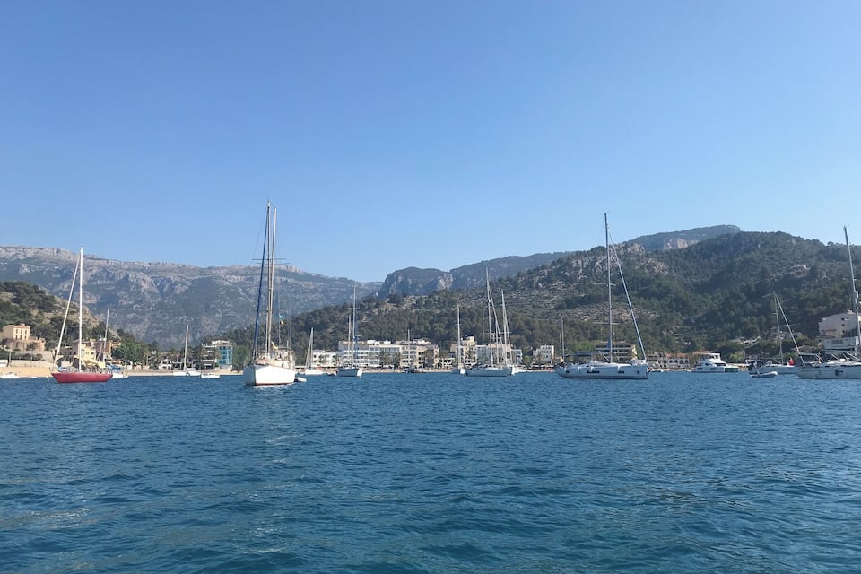 A marina with sailboats and yachts docked, a sandy beach, and a mountainous backdrop.