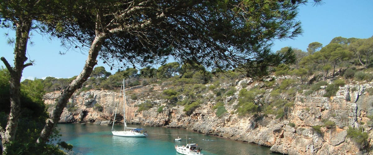 A clear turquoise bay with a sailboat and a motorboat anchored near rocky cliffs.