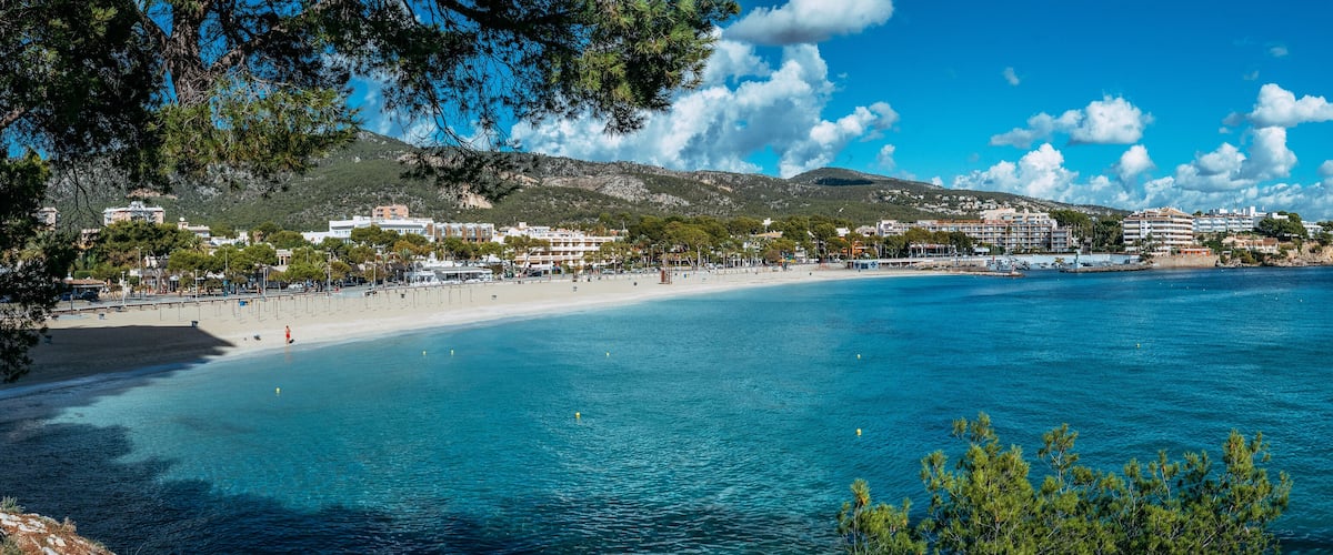 A coastal town with a sandy beach, clear blue waters, and a backdrop of hills and buildings.