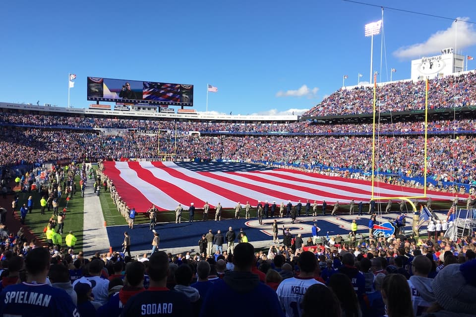 Um estádio lotado de espectadores, uma bandeira americana e uma tela grande exibindo um evento.