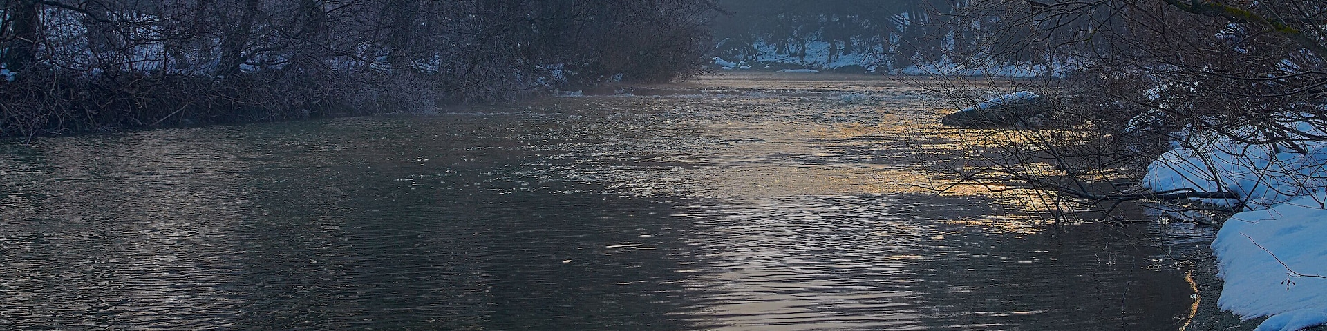 Un río con nieve en sus orillas y árboles desnudos al fondo.