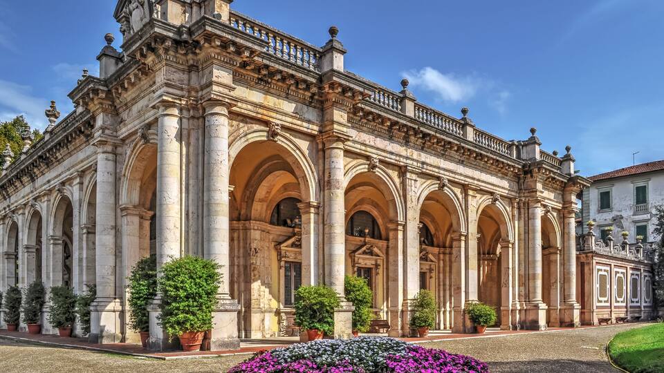 A historic building with ornate columns and arches, surrounded by potted plants and a flower bed.
