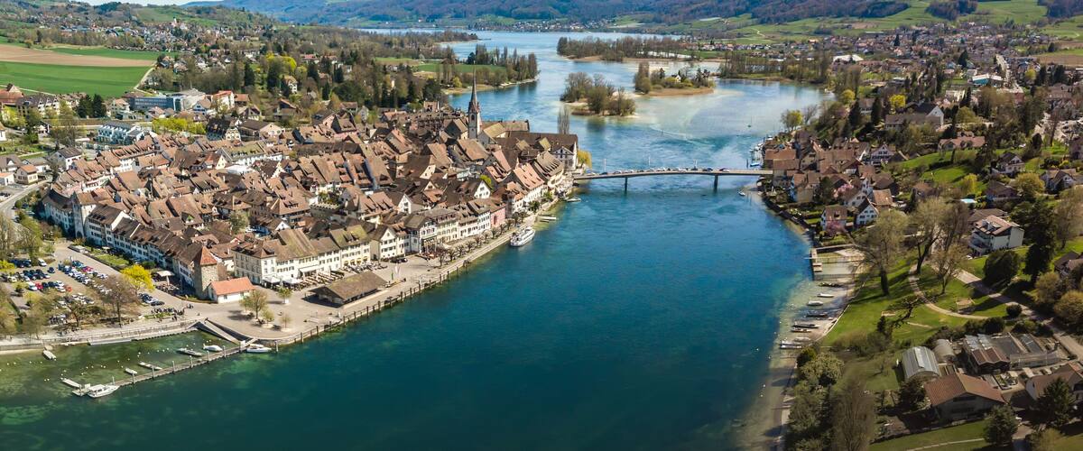 An aerial view of a riverside town with historic buildings, a bridge, and surrounding hills.