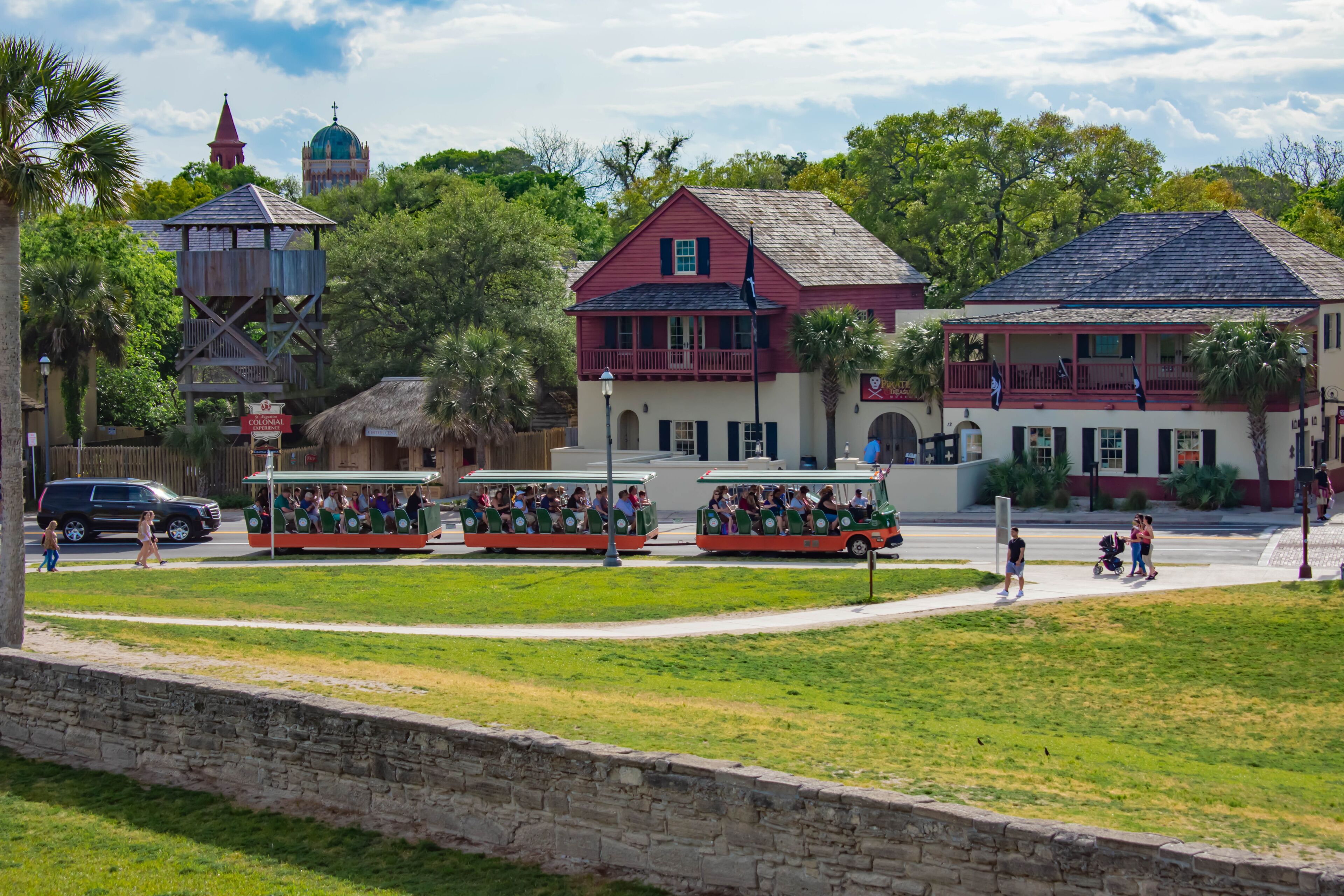 A colonial-style building with a red roof, a thatched-roof structure, and a green-domed church in the background.