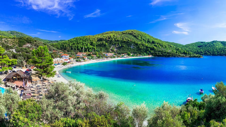 A coastal town with a turquoise bay, surrounded by lush green hills and a clear blue sky.