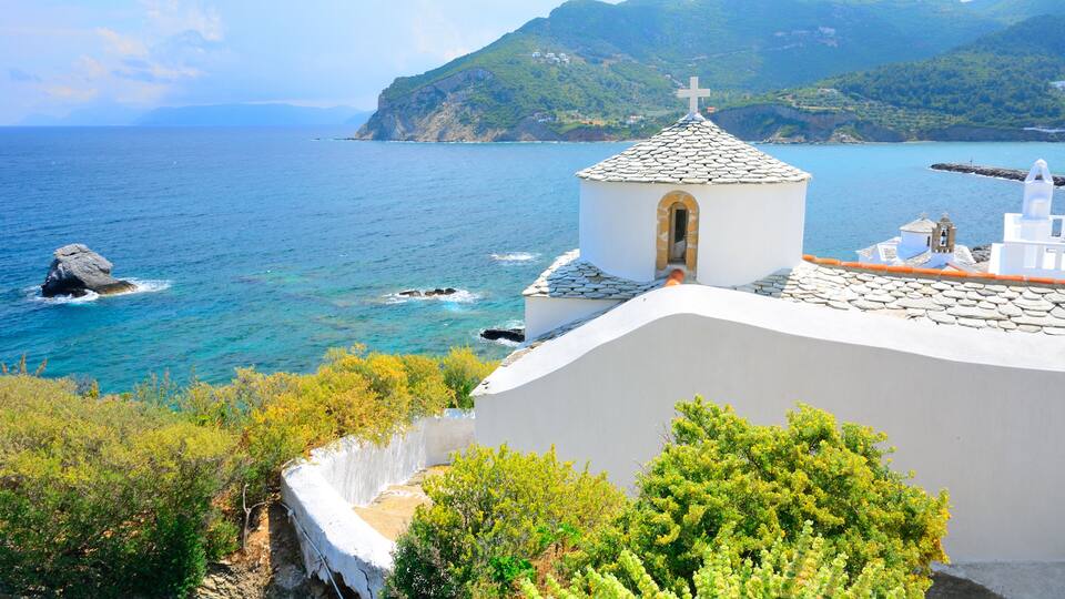 A coastal church with a cross on top, surrounded by greenery and overlooking the sea.