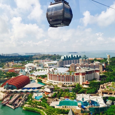 A cable car above a coastal resort with buildings, a marina, and a large dome structure.