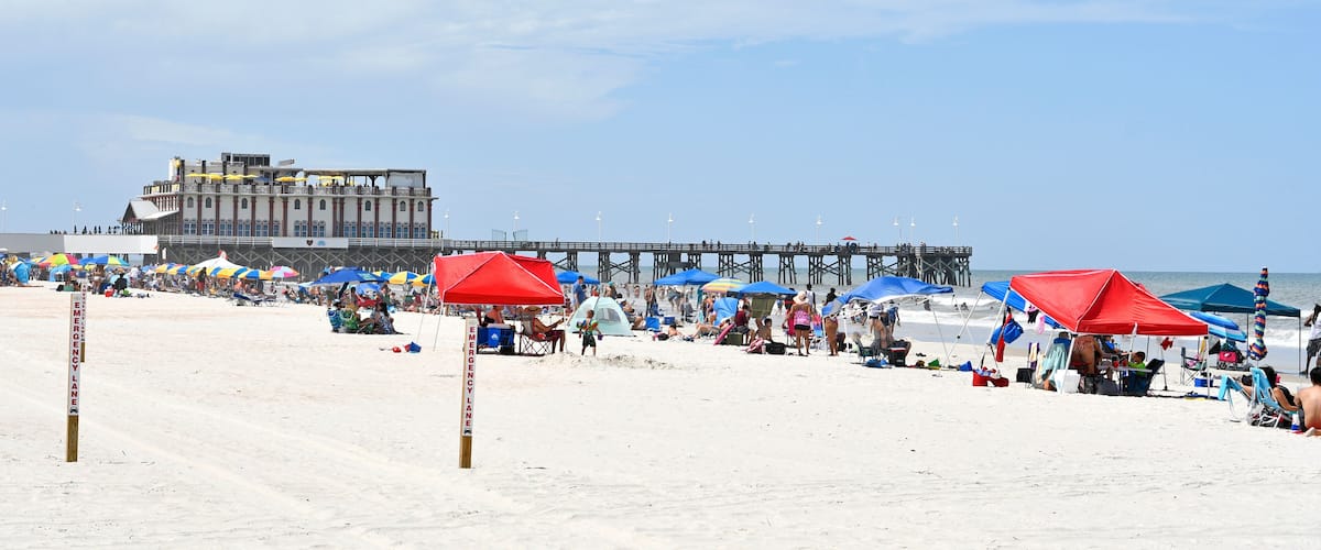 A crowded beach with umbrellas, a pier, and a large building in the background.