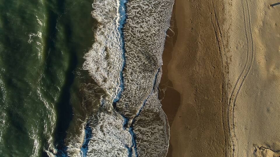 An aerial view of a beach with waves crashing onto the shore and a clear demarcation between the water and the sandy beach.