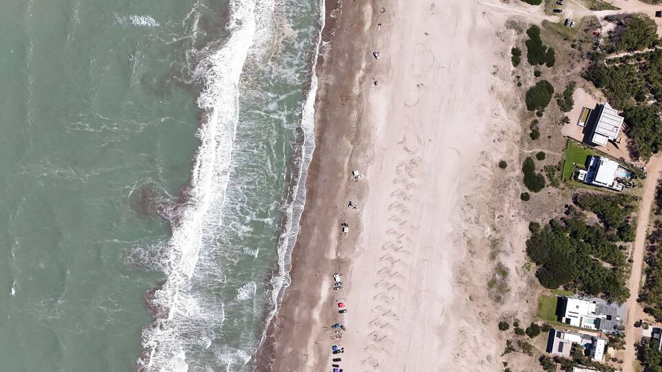 An aerial view of a beach with waves crashing onto the shore, a row of beach umbrellas, and several buildings in the background.