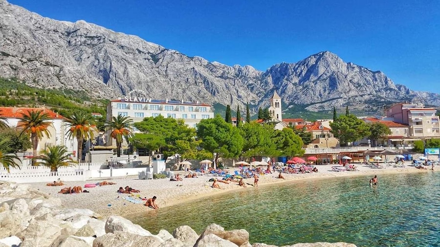 Un pueblo costero con playa, aguas azules cristalinas, montañas rocosas y edificios de tejados rojos.