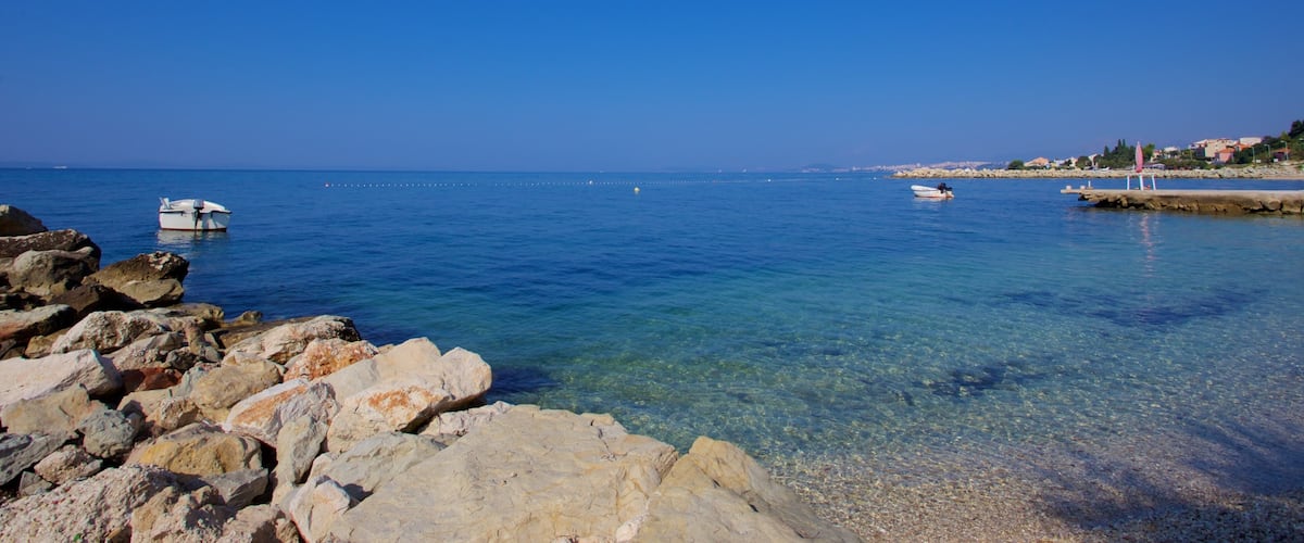 A clear blue sea with rocky shores, a small boat, and a distant shoreline with buildings.