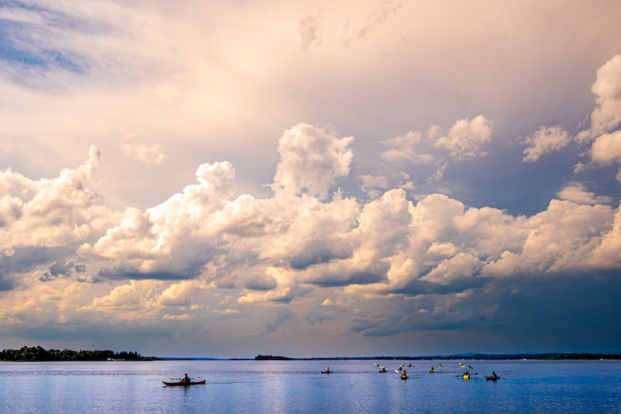 A calm lake with several kayaks and canoes, surrounded by a cloudy sky.